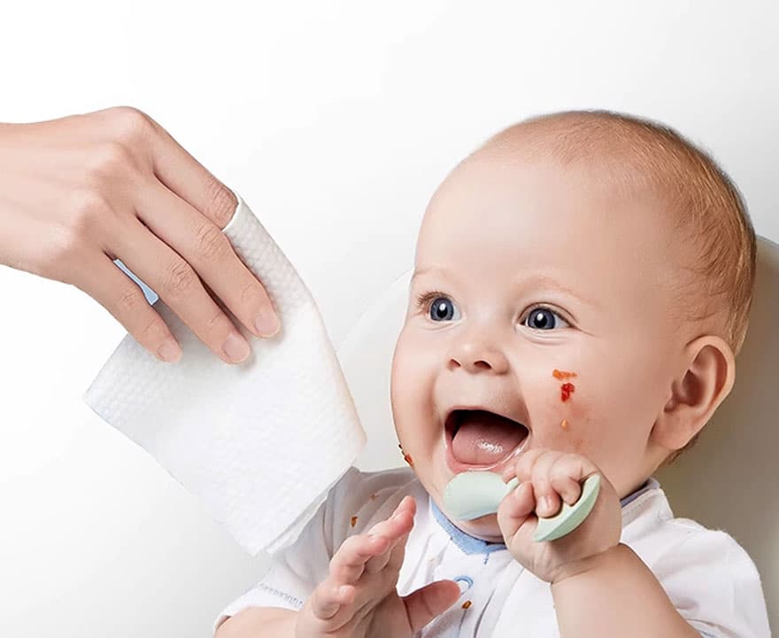 A mother's hand using a pearl-textured embossed wet wipe to gently clean a baby's messy face during feeding time, showcasing Sywipe's private label baby care wipes solutions.