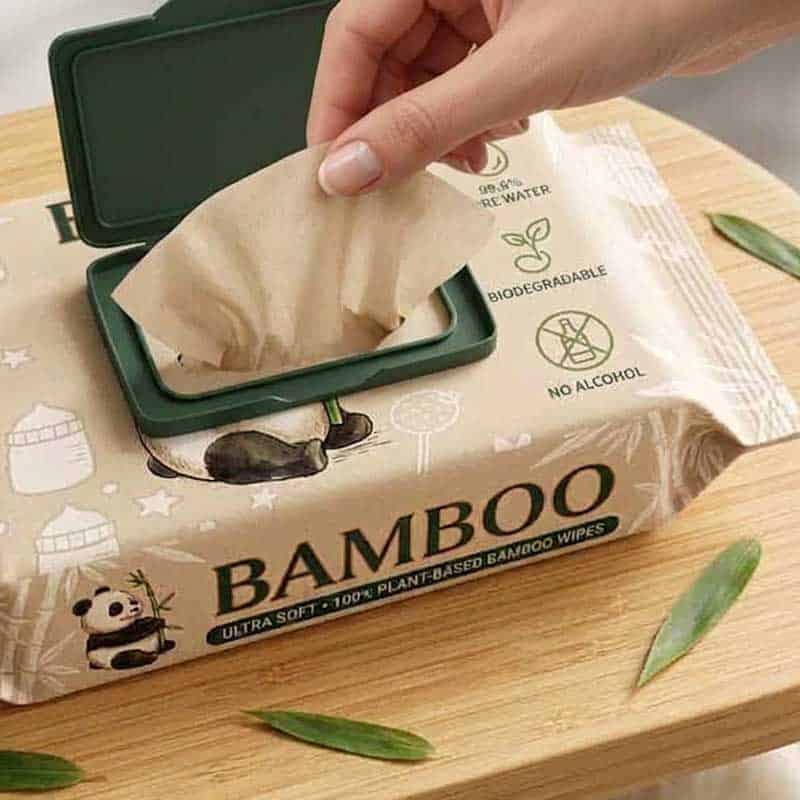 A hand pulling a natural unbleached biodegradable bamboo wet wipe from a flow pack with a green lid, featuring panda designs and bamboo leaves on a wooden table.