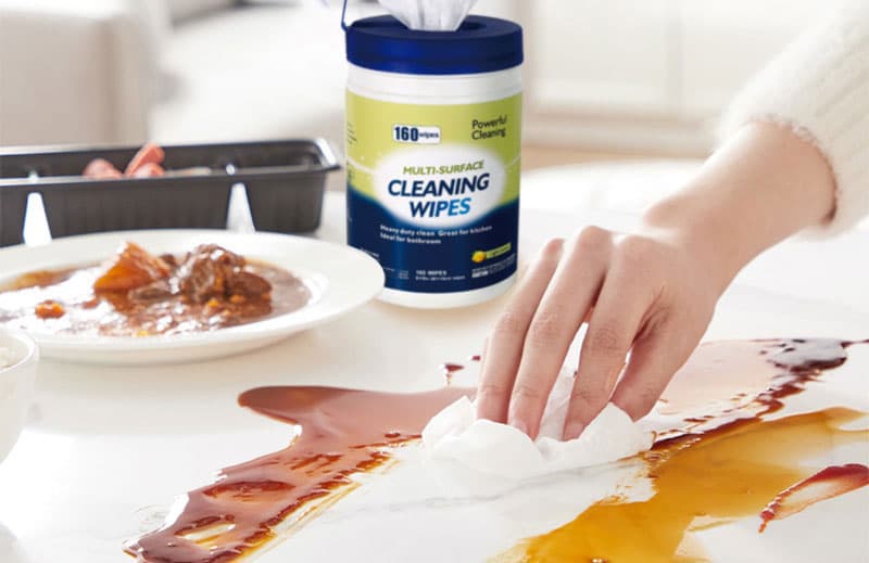 Hand wiping sauce stain on a white table using a 160-count canister of Sywipe multi-surface household cleaning wipes.